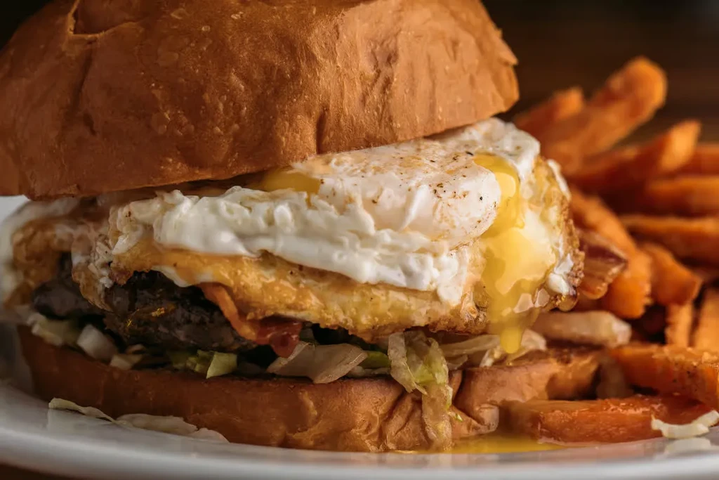 Tall hamburger with two fried eggs, bacon, sweet potato fries.