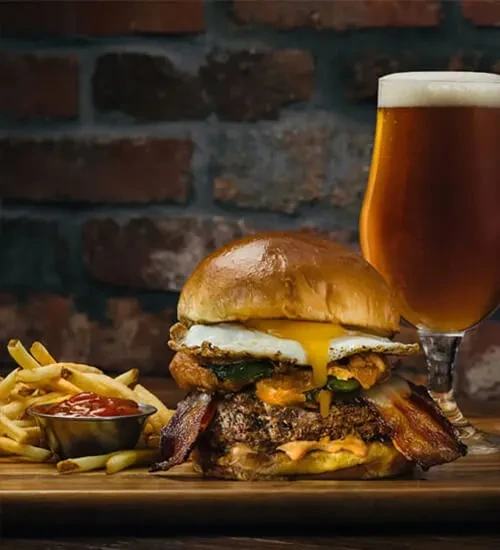 Stacked gourmet burger with runny yolk, fries, ketchup, and beer.