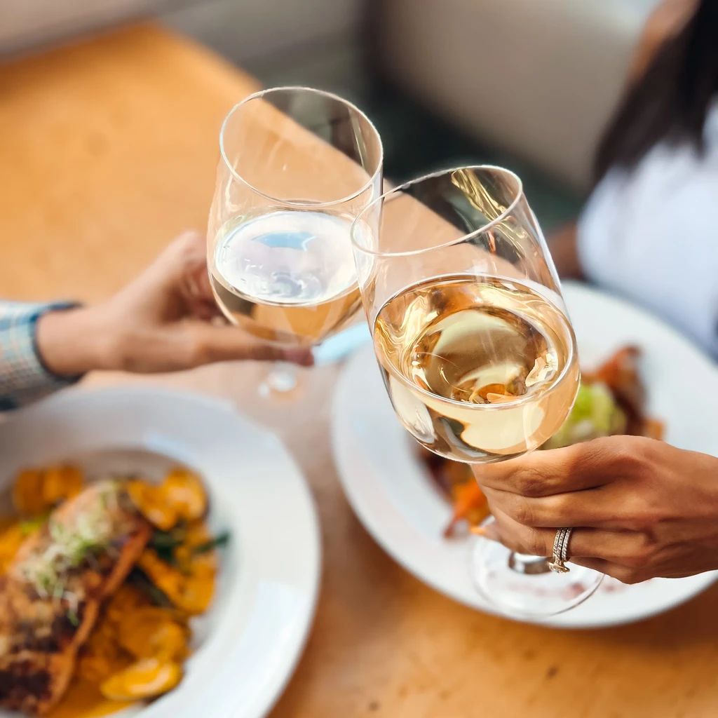 Two hands clinking white wine glasses over plates of food.