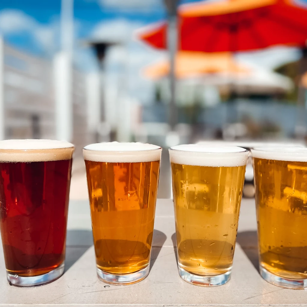 Four glasses of frothy beer, varied colors, lined up outdoors.