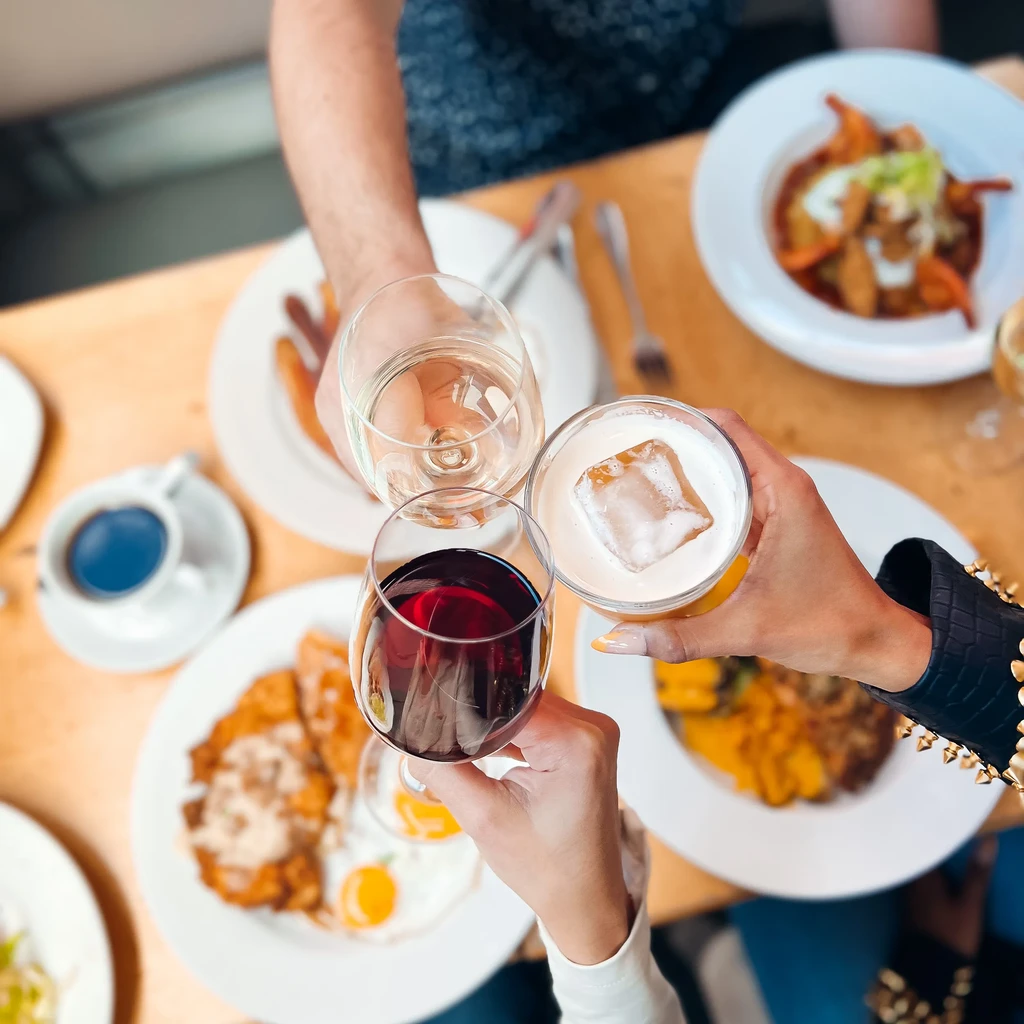 Four hands toast with wine and a cocktail over a table of food.