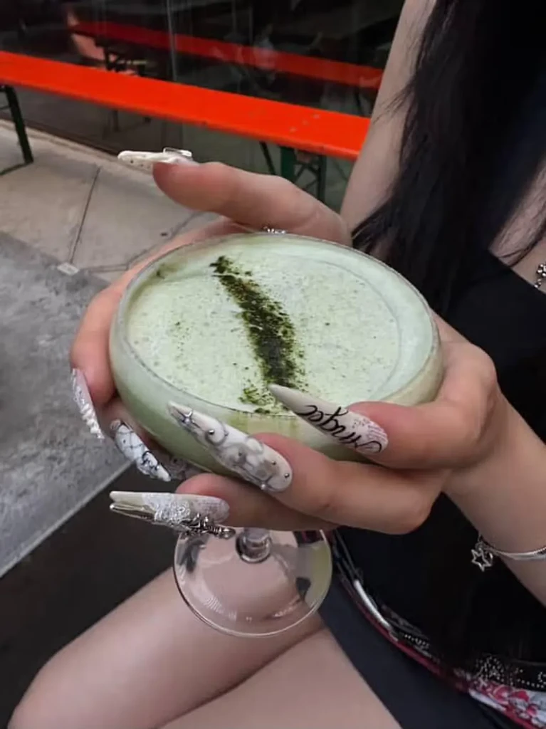 Hands with intricate stiletto nails holding a foamy green drink.