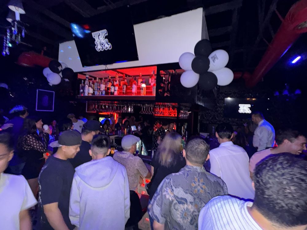 A dark, crowded bar with glowing screens, bottles, and balloons.