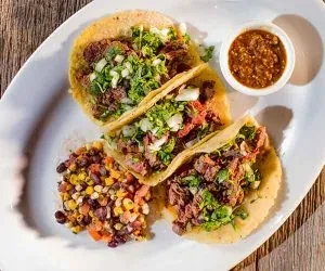 Three meat tacos with cilantro, onions, corn salad, and salsa.