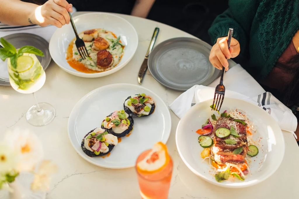Hands eating scallops, seafood, seared meat, and drinks on a table.