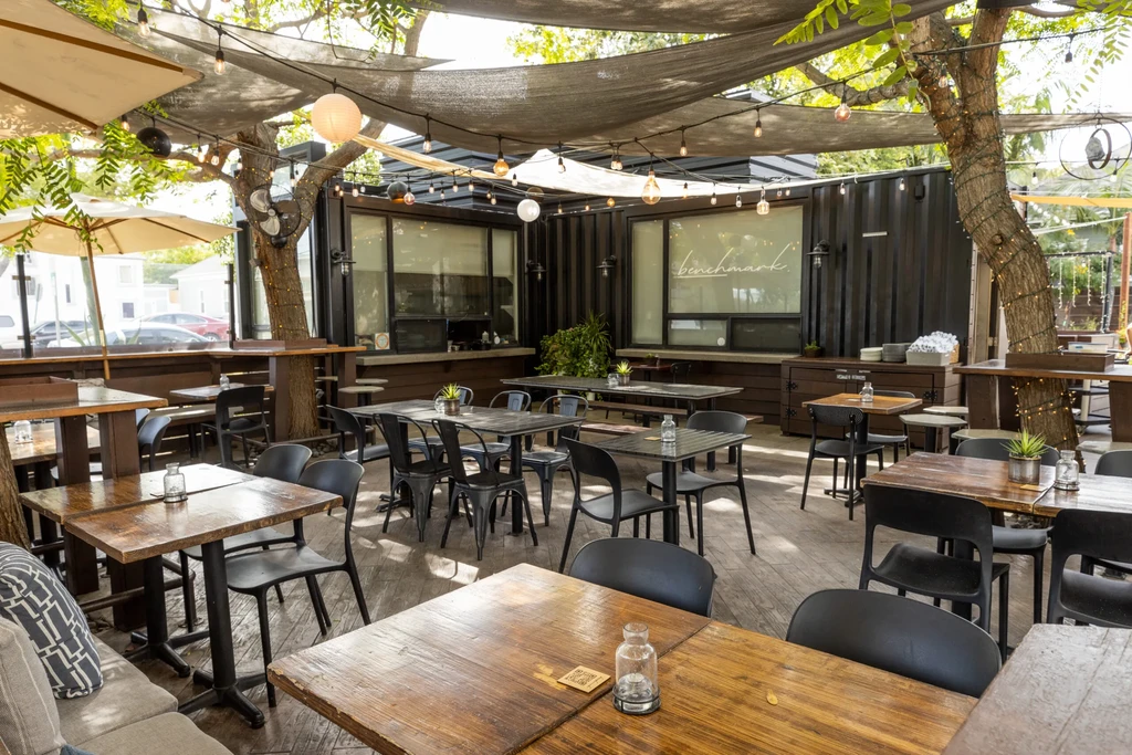 Shaded outdoor dining area with tables, string lights, trees, and "benchmark" sign.