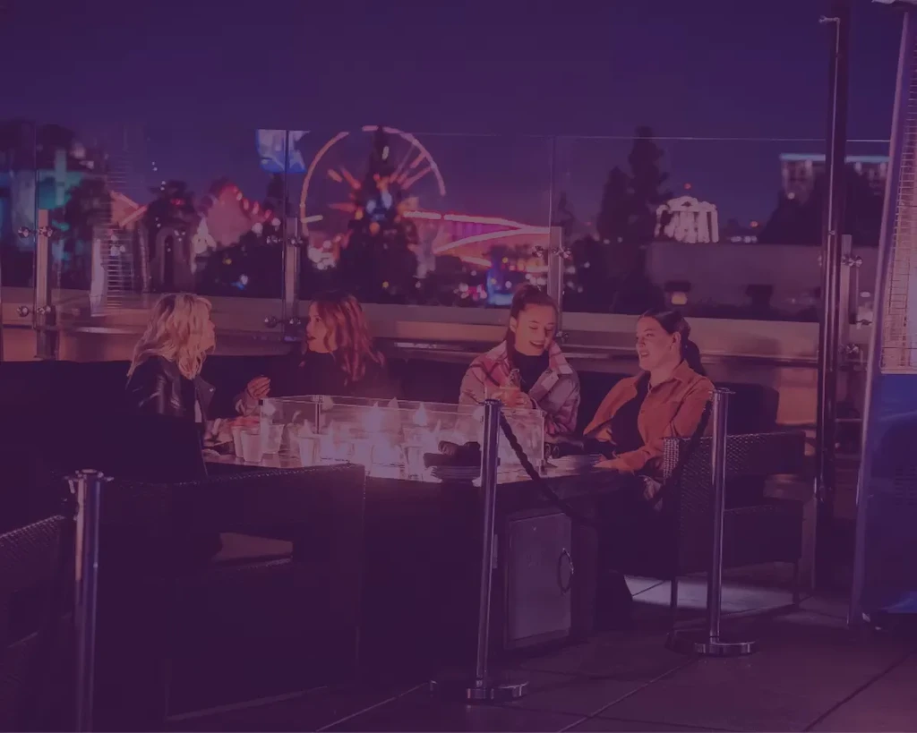 Nighttime patio: four women by fire pit, city skyline, lit Ferris wheel.