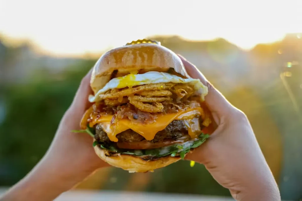 Hands hold a gourmet burger with beef, cheese, egg, and onion rings.