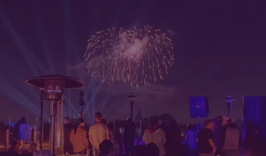 Crowd watches purple fireworks and a glowing patio heater at night.