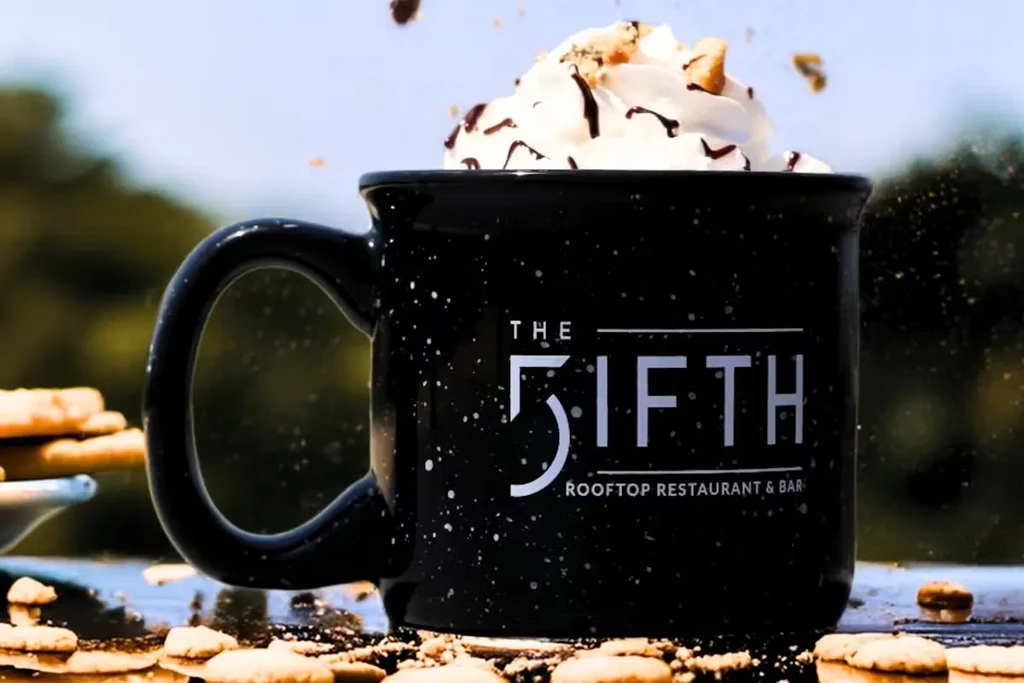 THE 5IFTH branded black mug, whipped cream drink with chocolate, outdoors.