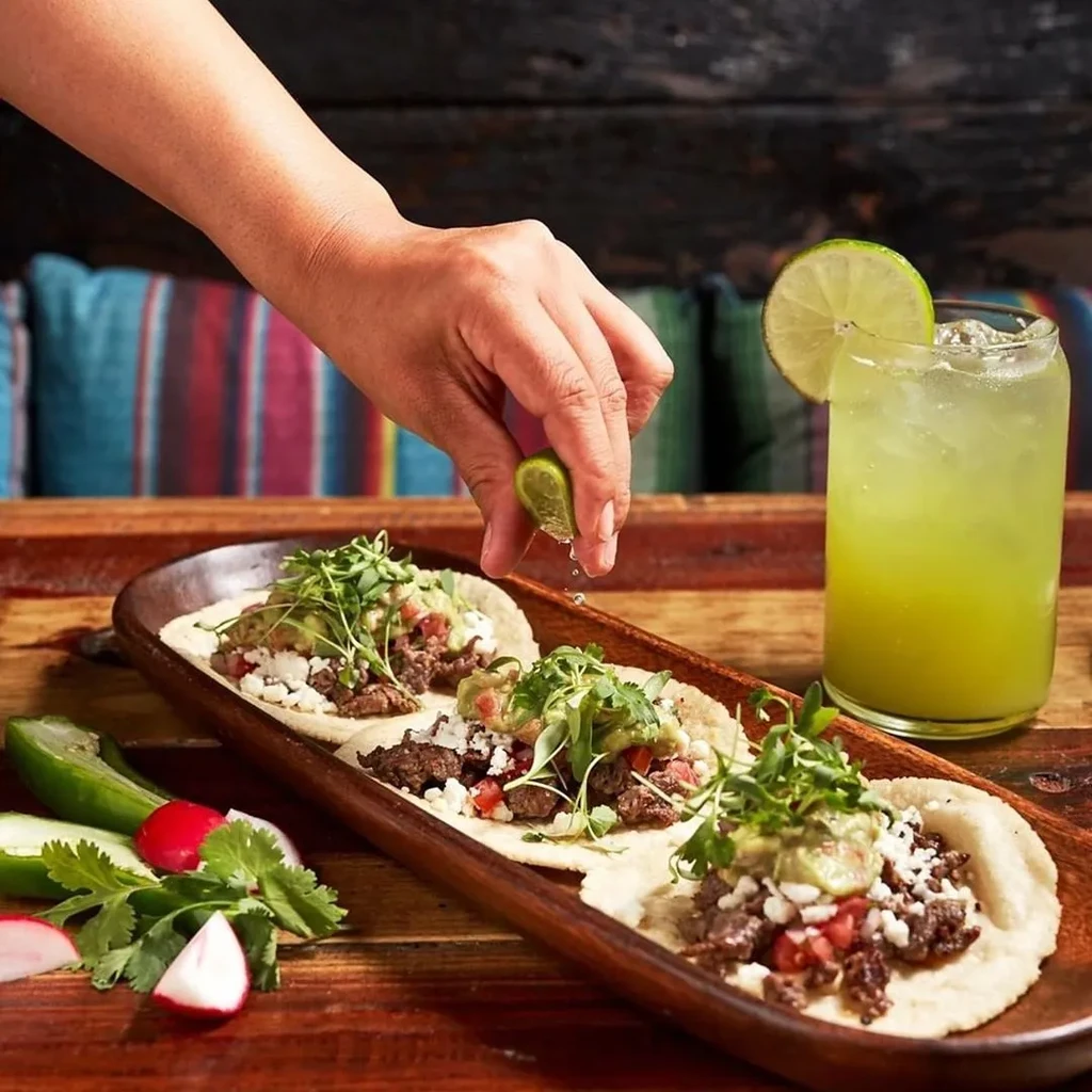 Hand squeezes lime over three tacos on dark wooden tray.