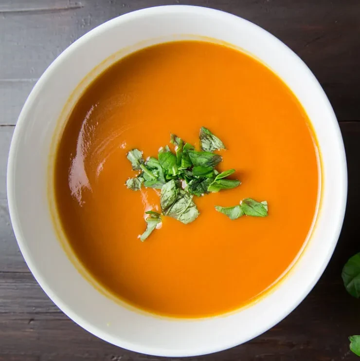 Vibrant orange soup in a white bowl, garnished with fresh green herbs.