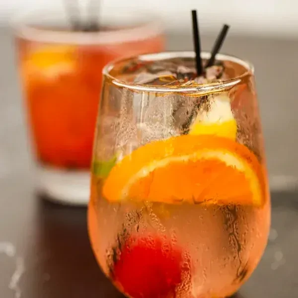 Two cold drinks with ice, citrus, and a cherry on a dark surface.