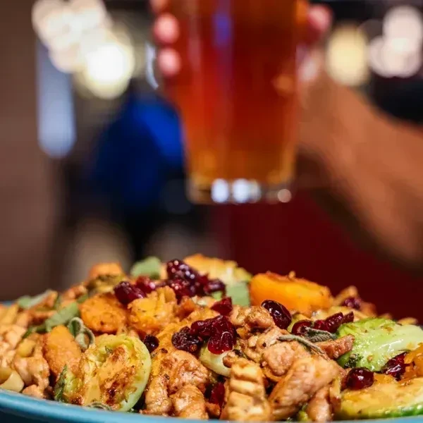 Roasted chicken, vegetables, cranberries on plate; blurred hand holds a beer.
