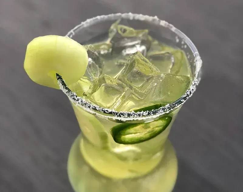 Green cocktail with salted rim, ice, jalapeño, and melon ball garnish.