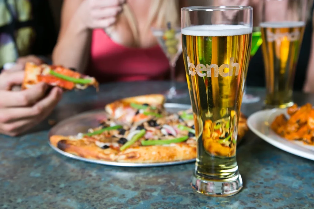 Golden beer glass etched 'bench,' with pizza and wings in background.