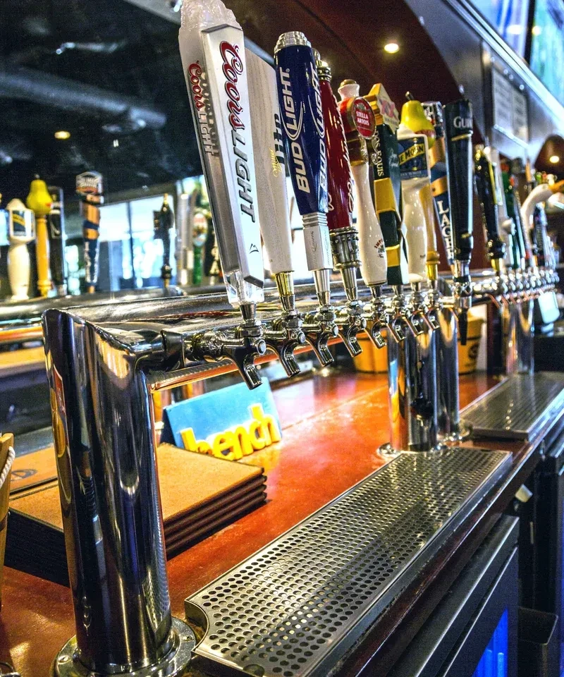 Variety of chrome beer taps on a warm wooden bar counter.