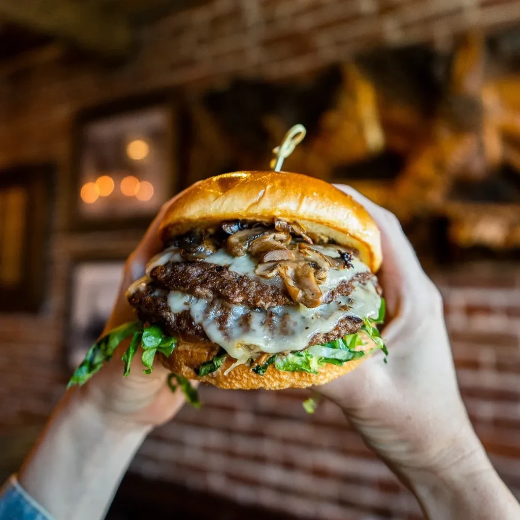 Two hands hold a large double-patty burger with cheese and mushrooms.