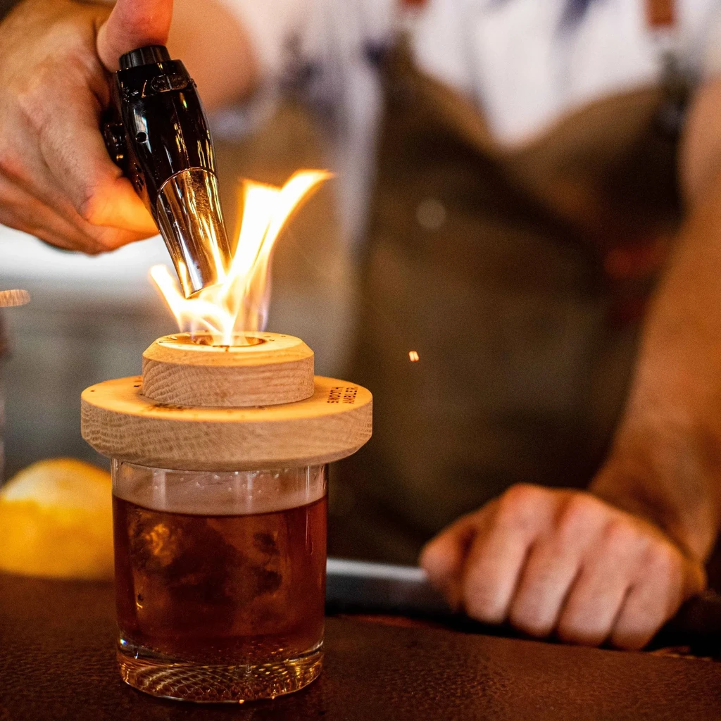 Close-up of a cocktail flaming on a wooden device at a bar.