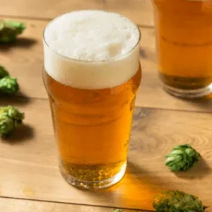 Pint glass of golden beer, condensation, and green hops on wood.