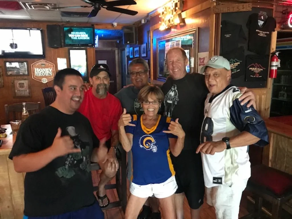 Six smiling people in a rustic bar, two making shaka gestures.