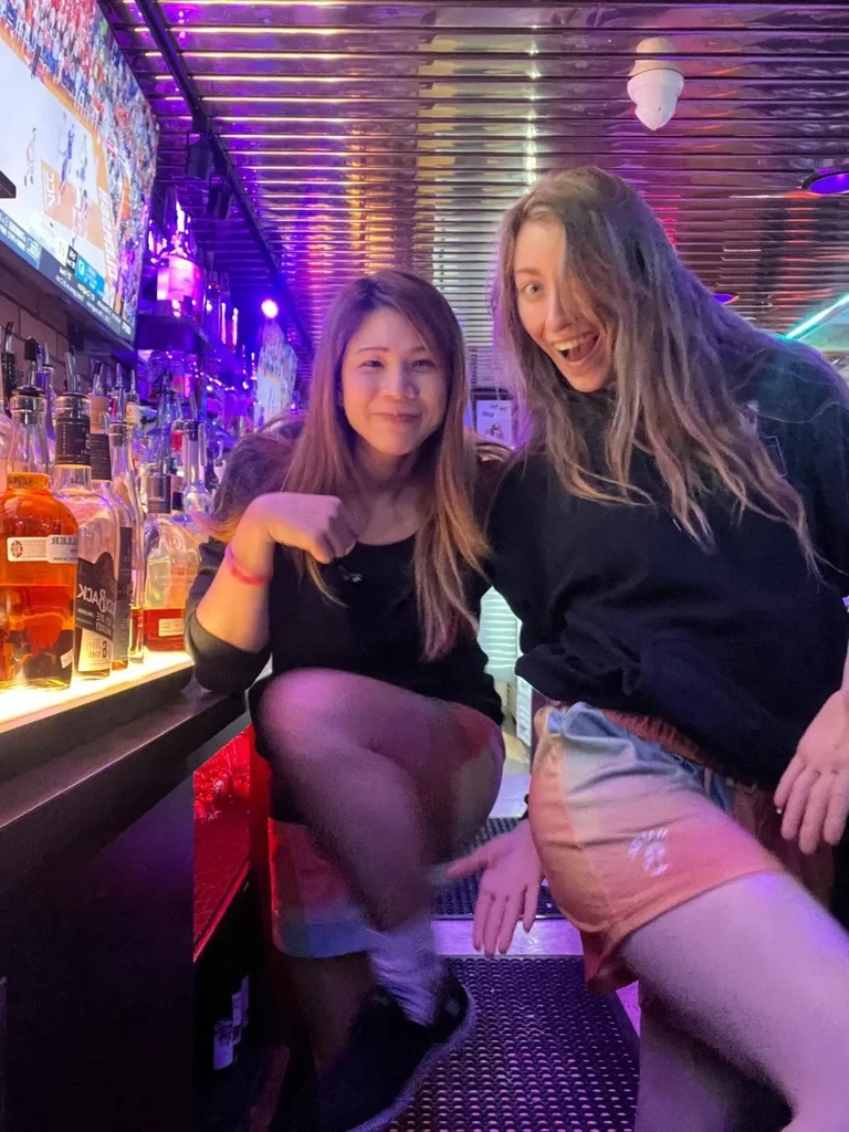 Two women smiling and laughing at a neon-lit bar counter.