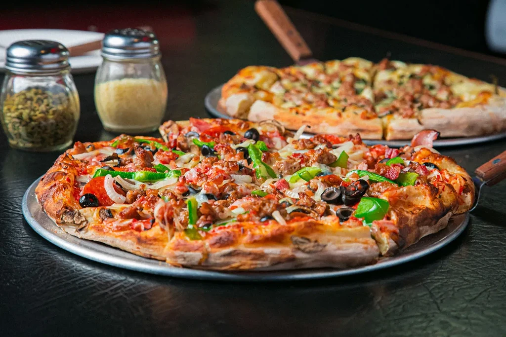 Supreme pizza with lifted slice, a second pizza, and spice shakers.