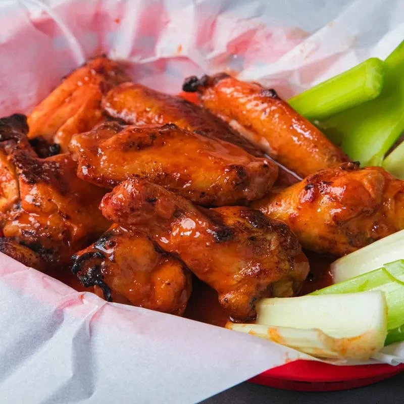 Generously sauced, charred buffalo wings and celery in a red basket.