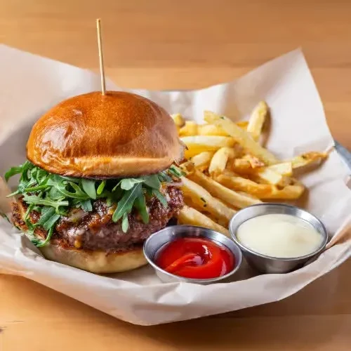 Arugula burger and golden french fries with sauces on a wooden surface.