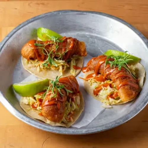 Three crispy fish tacos with red sauce, microgreens, and lime.