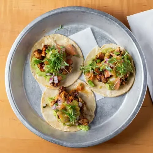 Three chicken tacos with guacamole and salsa on a metal tray.
