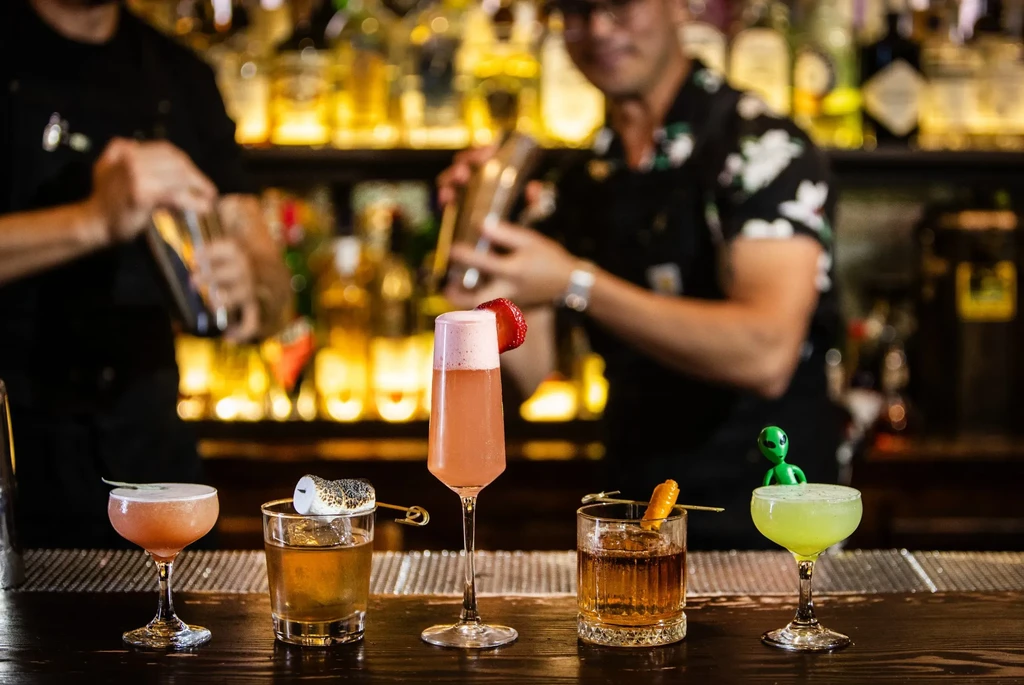 Five distinct garnished cocktails on a bar counter with blurred bartenders.