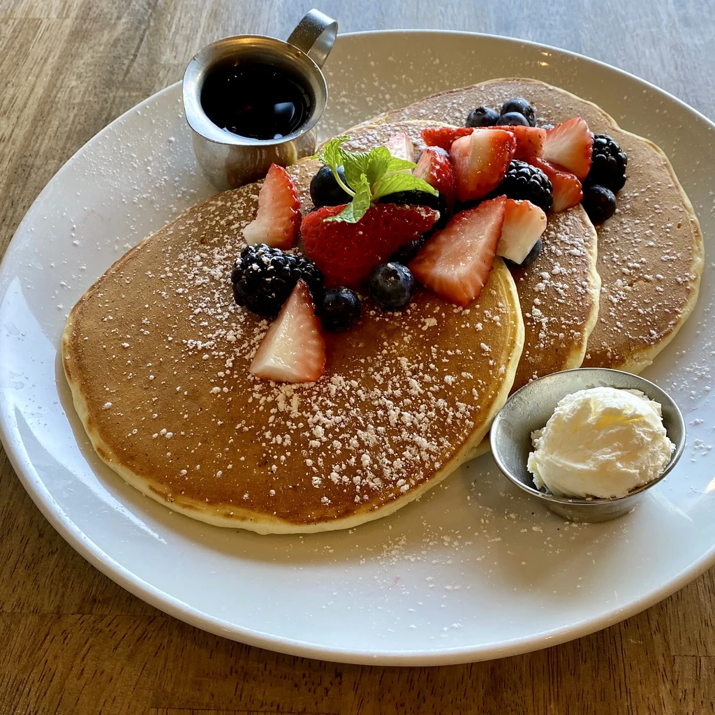 Three fluffy pancakes with berries, mint, powdered sugar, syrup, and cream.