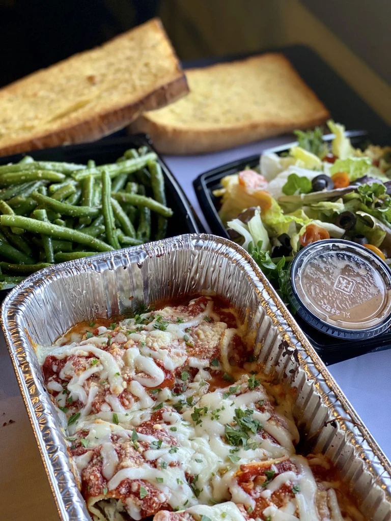 Baked pasta with cheese, green beans, salad, and garlic bread in takeout containers.
