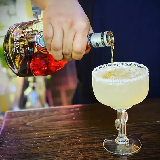 Hand pours amber liquid from bottle into salt-rimmed cocktail glass.