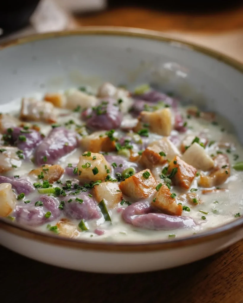 Rustic bowl of creamy purple gnocchi, seared pieces, and green herbs.