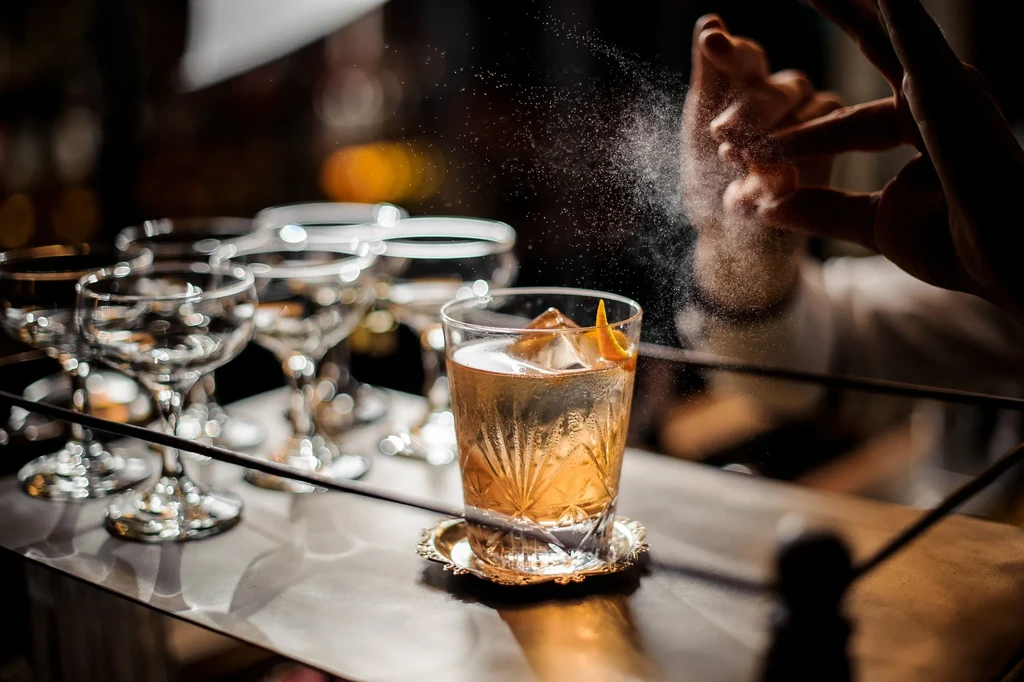 Bartender sprays mist over a crystal glass cocktail with orange peel garnish.
