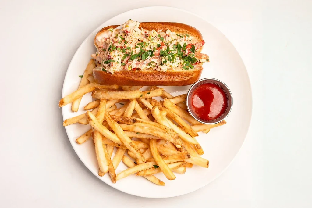 Lobster roll, golden french fries, and ketchup on a white plate.