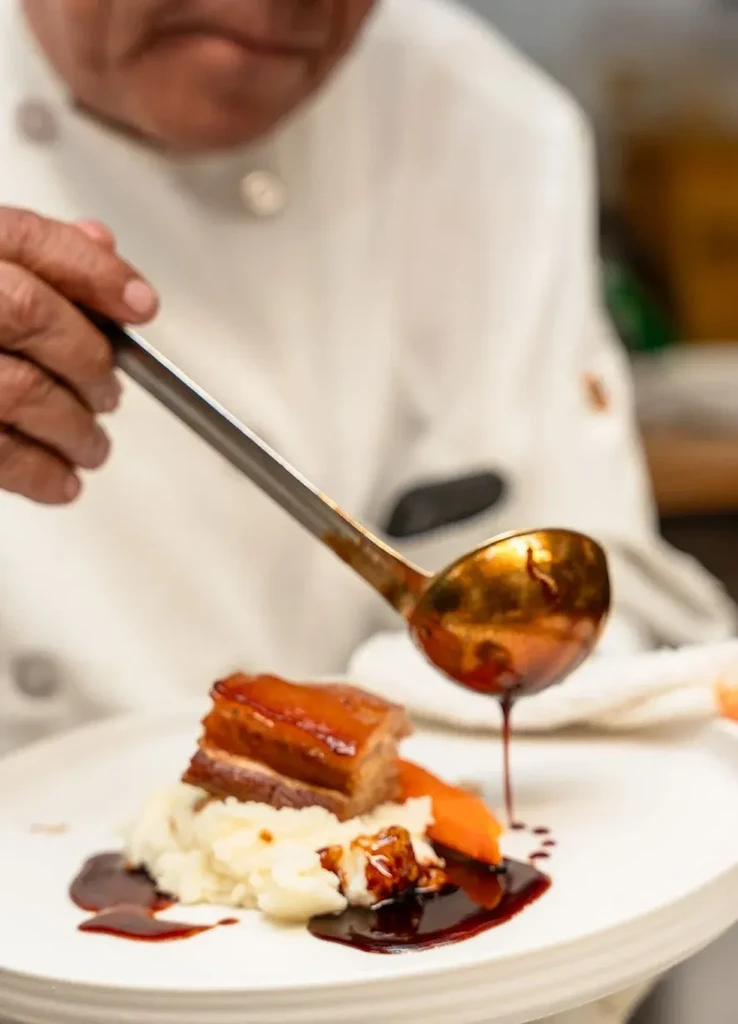 Chef's hands plate pork belly, potatoes, orange vegetable, and dark sauce.