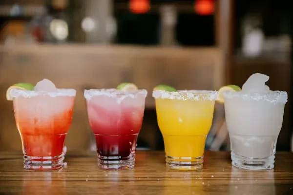 Four colorful, salt-rimmed drinks garnished with lime on a bar.