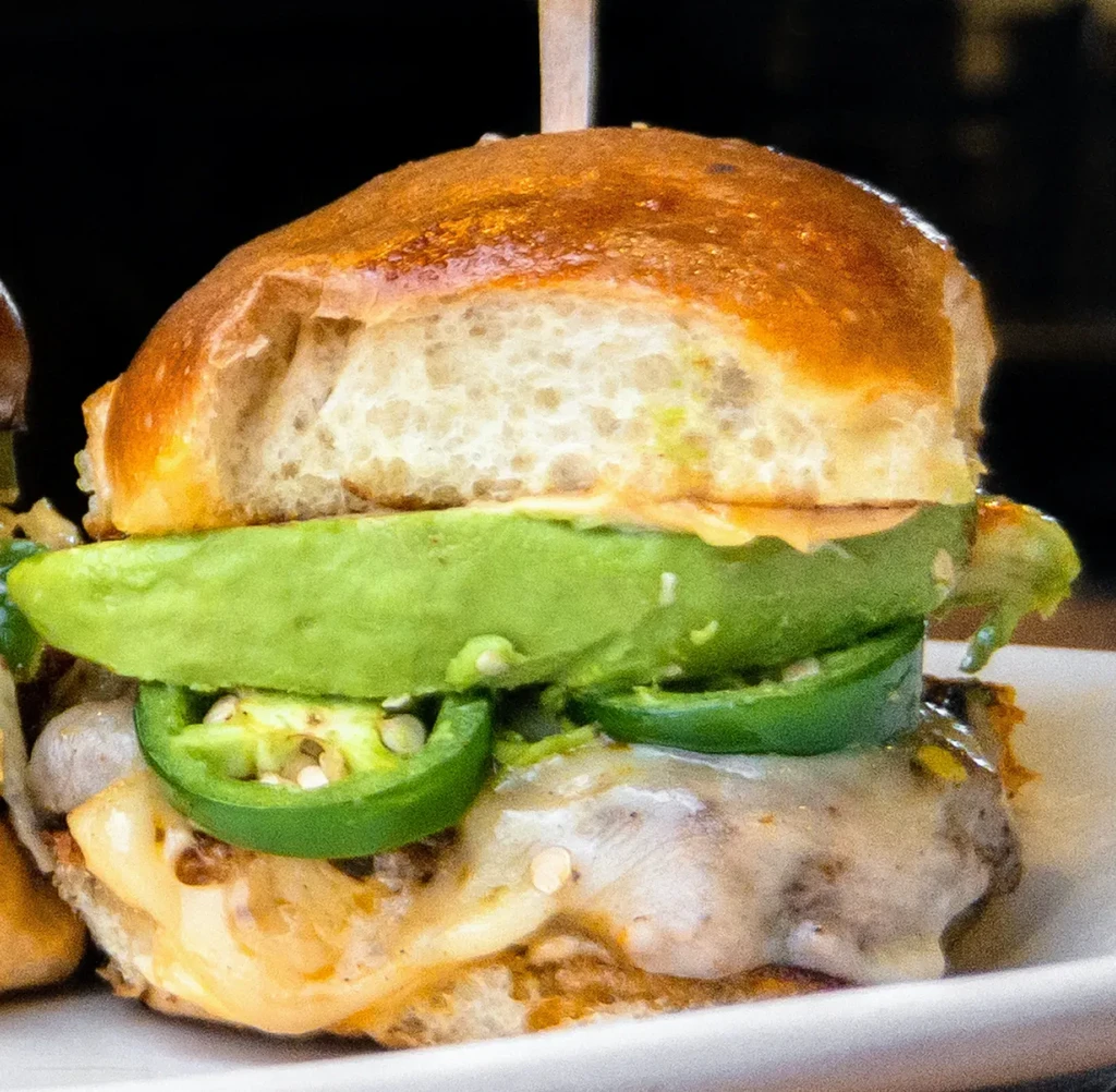 Gourmet slider with avocado, jalapeños, cheese, and a wooden skewer.