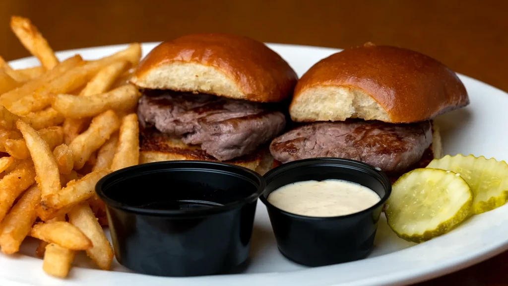 Two sliders with fries, sauces, and pickles on a white plate.