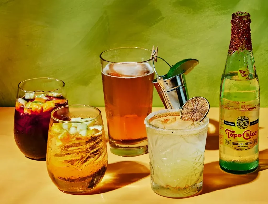 Five beverages, including beer and Topo Chico, on a peach surface.
