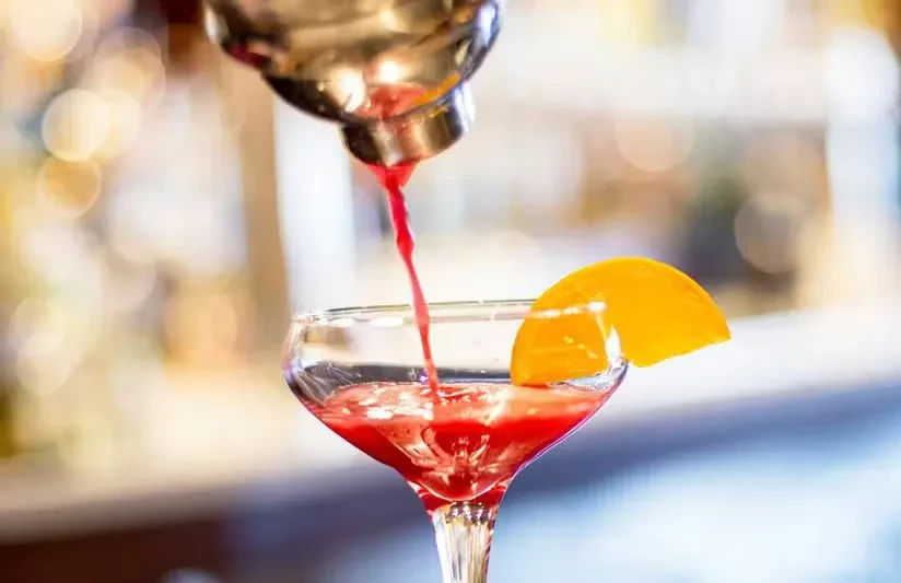 Cocktail shaker pouring red drink into a coupe glass.