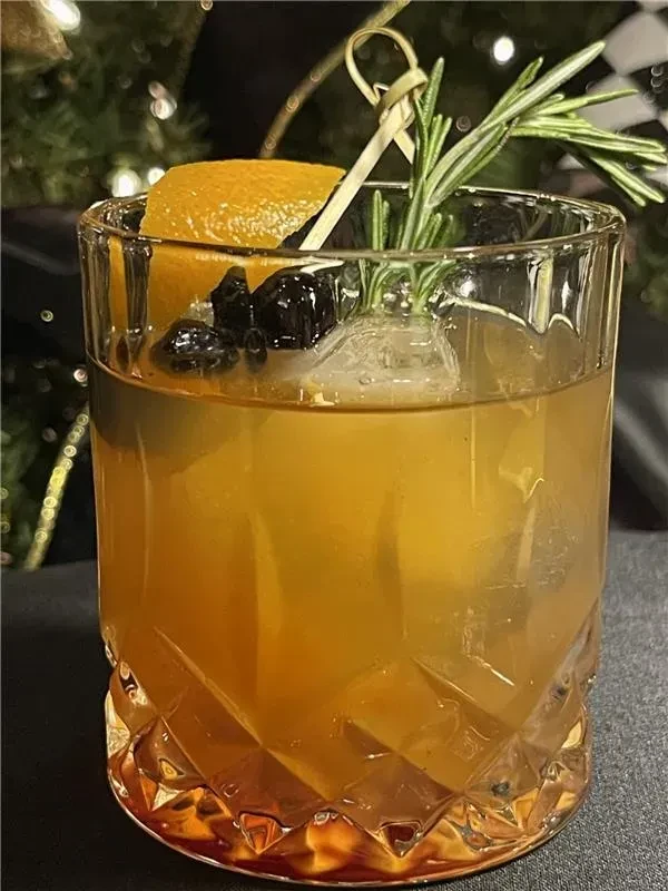 Cocktail in diamond-cut glass, garnished with orange, berries, and rosemary.
