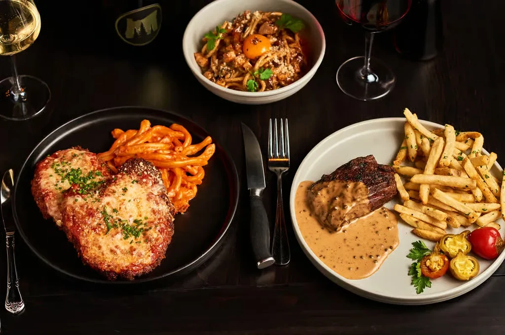 Flat lay of a table with chicken parmesan, steak, pasta, and wine.