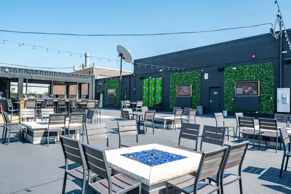 Outdoor restaurant patio with fire pit tables, bar, and TVs.