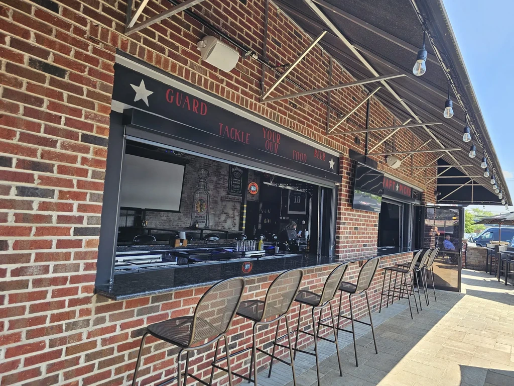 Outdoor bar with brick counter, metal stools, and a projection screen.
