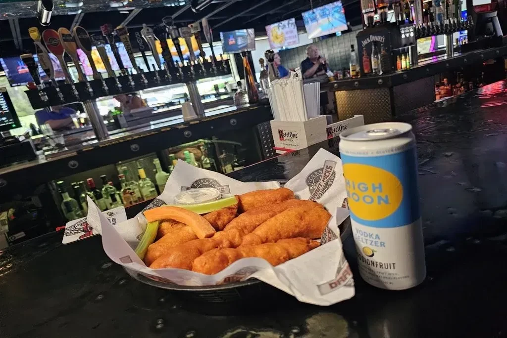 Fried chicken tenders, vegetables, dipping sauce, and seltzer at a bar.