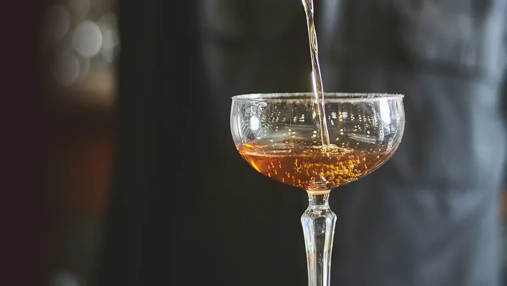 Amber liquid pours into a sugared coupe glass with bokeh background.